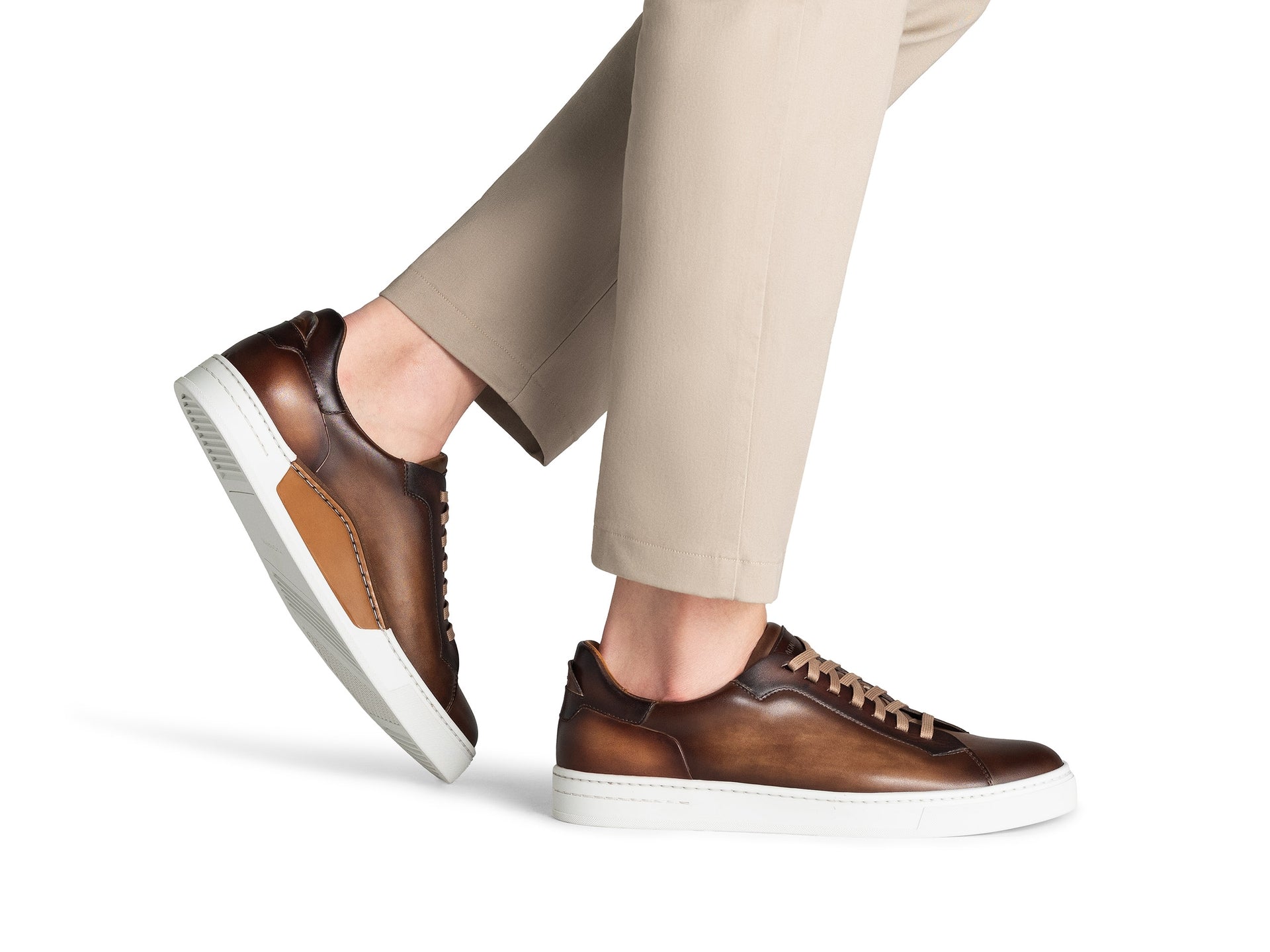 A person wearing tan pants and Magnanni Amadeo Sneaker by Magnanni in brown leather with white soles, standing against a white background.