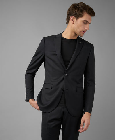 A man in a dark suit and black shirt stands against a gray background, looking down and to his right with one hand in his pocket, effortlessly elevating his style with the Robert Barakett Georgia Crew 23336 tee.