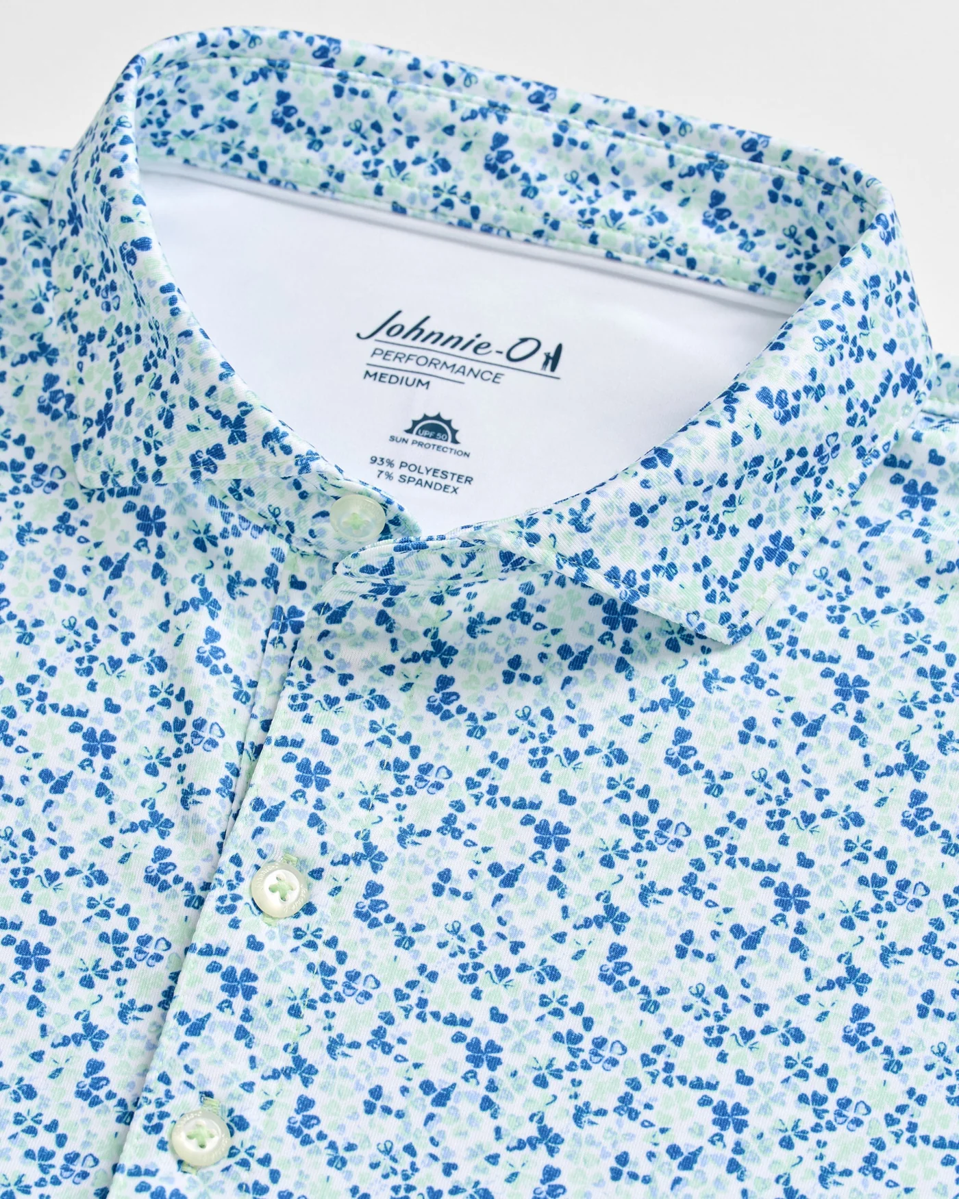 Close-up of the Johnnie-O Charmer Jersey Polo featuring a blue and green floral pattern, white buttons, and a Johnnie-O Performance Medium label inside the collar.