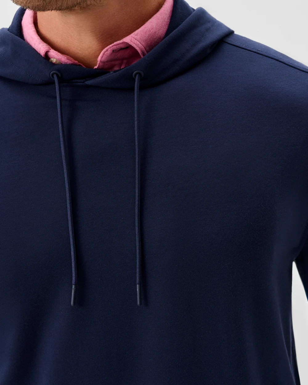 A person wears the Johnnie-O Willard Performance Hoodie Big & Tall in navy blue—an athletic-fit piece from Johnnie-O—layered over a pink collared shirt, with only their lower face, neck, and upper chest visible.
