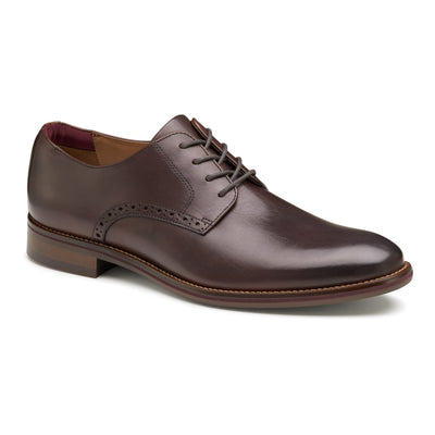 The Johnston & Murphy Conard 2.0 Plain is a brown leather dress shoe with laces, low heel, decorative side perforations, a slightly rounded toe, and cushioned footbed for comfort, shown on a white background.