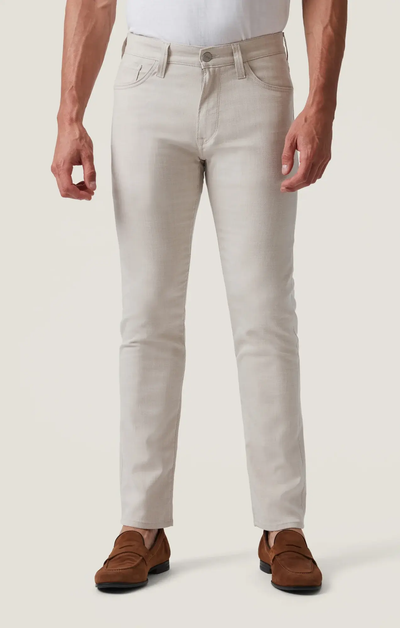 A man wears the 34 Heritage Courage Cross Twill slim-fit jeans in light beige, crafted from breathable twill fabric by 34 Heritage, paired with a white tucked-in shirt and brown suede loafers, standing against a plain off-white background.