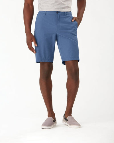 A man wearing a light gray shirt, Tommy Bahama Chip Shot Shorts Big & Tall in blue, and gray slip-on shoes stands with one hand in his pocket against a plain white background.