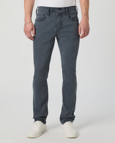 A person wears Paige Federal Slim Straight Jeans by Paige in dark gray, styled with a white shirt and white sneakers, standing against a plain light background.