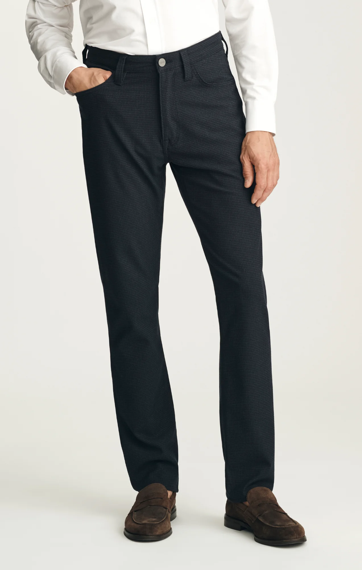 Wearing the 34 Heritage Courage Supreme Plaid pants by 34 Heritage, a person stands with one hand in their pocket against a plain background, paired with a white long-sleeve shirt and brown suede shoes.