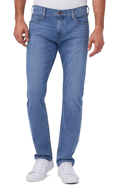 A man wearing Paige Federal Straight Slim Jeans by Paige in a medium wash, paired with a white shirt and white sneakers, stands against a plain background.
