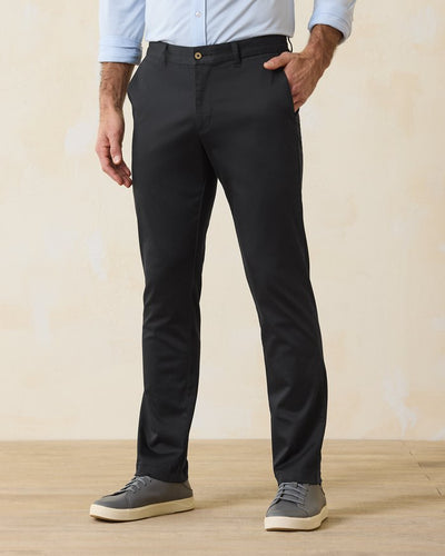 A man stands on a wooden floor wearing Tommy Bahama Boracay Pant Big & Tall chinos in black, a light blue long-sleeve shirt, and gray sneakers. His left hand in his pocket highlights this effortless Tommy Bahama look crafted with sustainable materials.