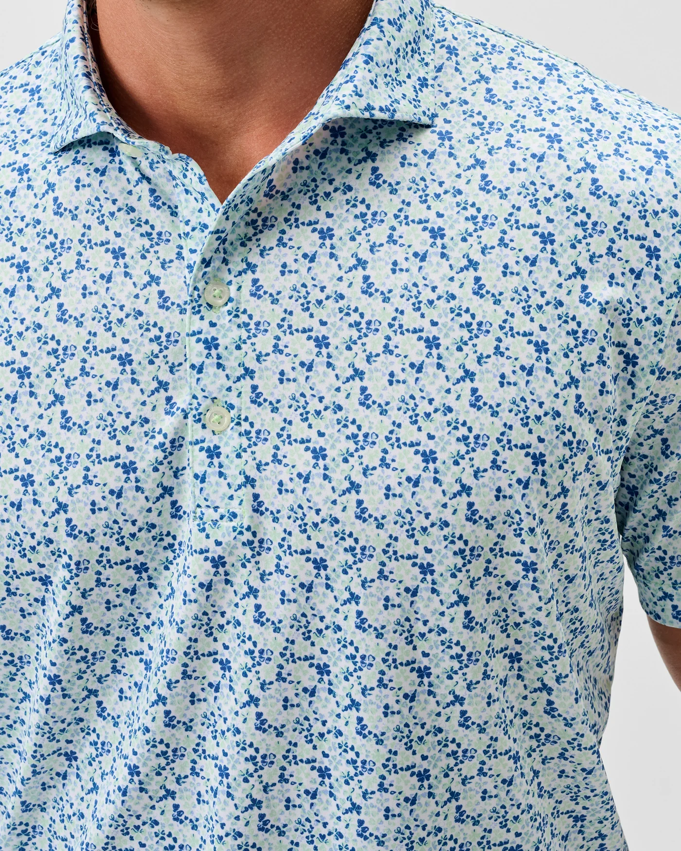 A person wearing the Johnnie-O Charmer Jersey Polo by Johnnie-O, featuring a light blue and white floral pattern with moisture-wicking fabric, shown from shoulders to mid-chest.