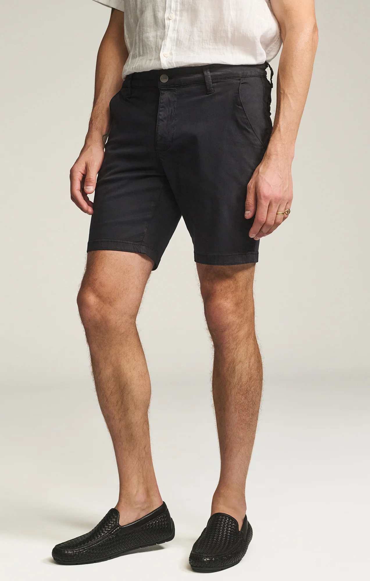 A man stands against a plain background wearing a white short-sleeve shirt, black 34 Heritage Arizona Twill Shorts by 34 Heritage, made from a cotton-Tencel blend, and black woven slip-on shoes.