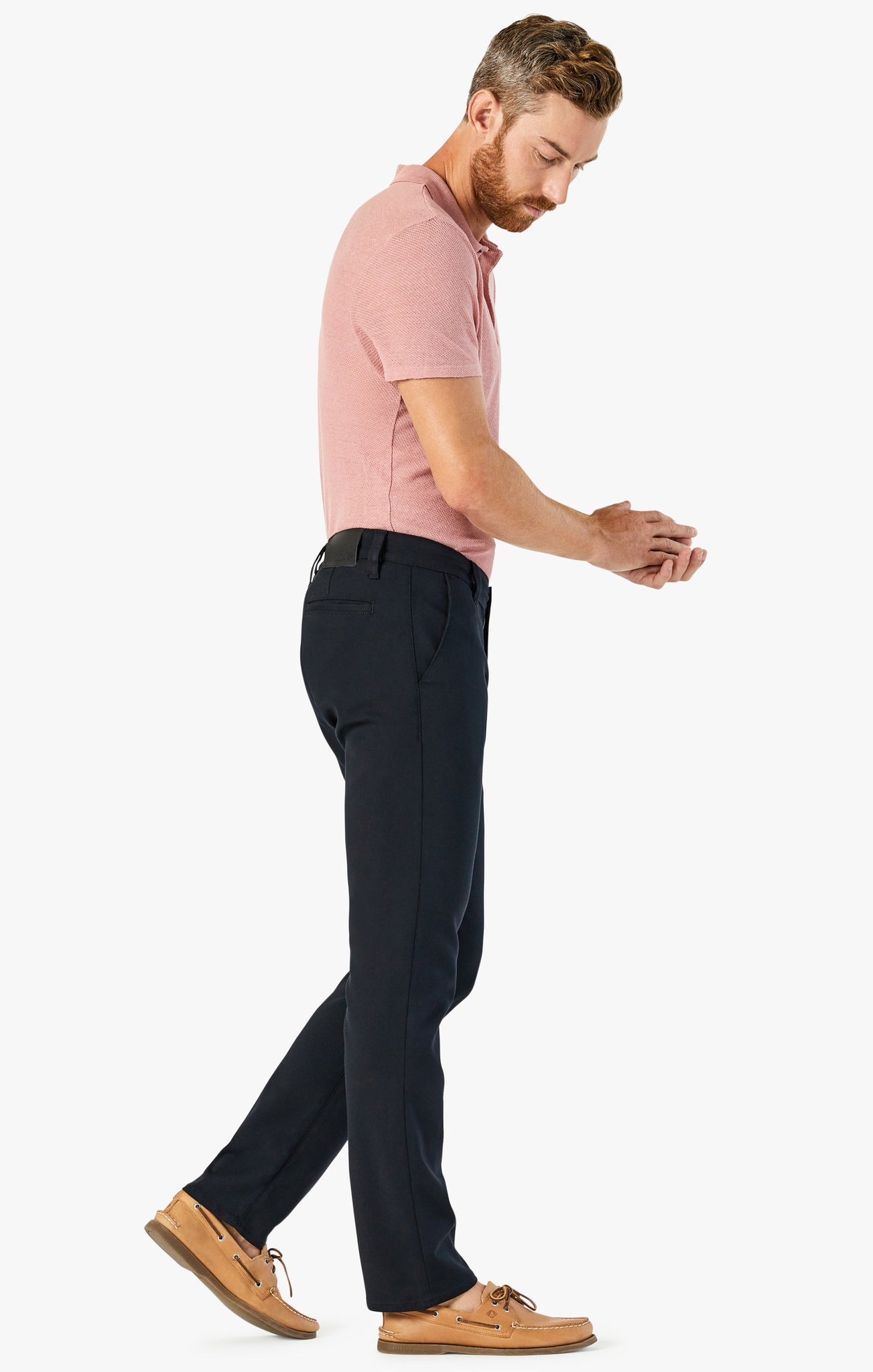A man with a beard, wearing a pink polo shirt, 34 Heritage Verona Pant High Flyer by 34 Heritage, and tan boat shoes stands against a white background, looking down at his hands.