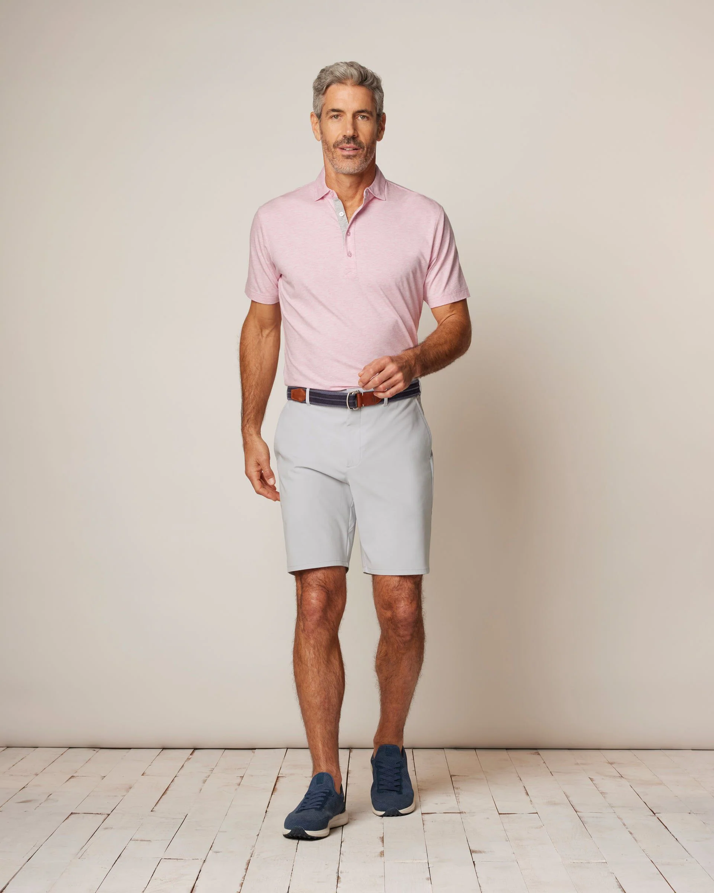 A man stands indoors against a neutral background wearing a light pink polo, Johnnie-O Mulligan Big & Tall Shorts by Johnnie-O, a belt, and navy sneakers.