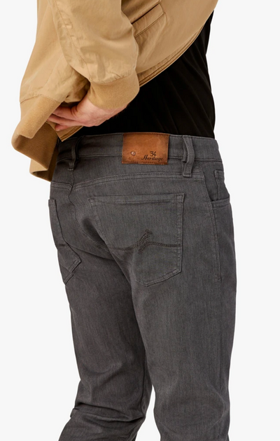 Person wearing 34 Heritage Courage Diagonal Pants in a straight fit gray style, paired with a tan jacket, viewed from behind. The pants feature a brown branded 34 Heritage leather patch on the waistband.