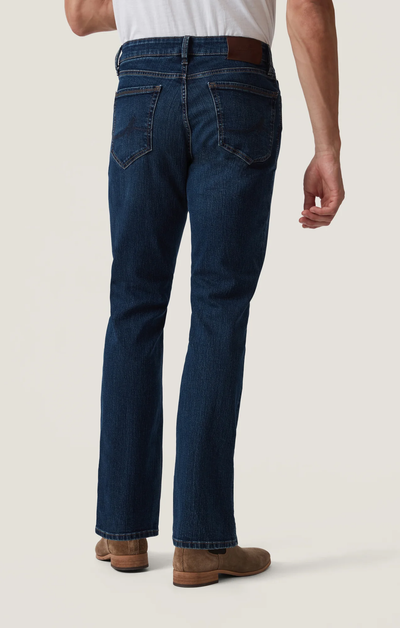 A man wearing 34 Heritage Charisma Mid Comfort jeans, a white shirt, and brown shoes stands facing away from the camera against a plain background.