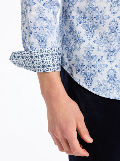 A person wears the Robert Graham Delancey Woven L/S Shirt B&T, a men’s cotton shirt by Robert Graham, featuring a blue allover floral print with striking details, turned-up sleeve cuffs revealing a contrasting geometric design, paired with dark pants.