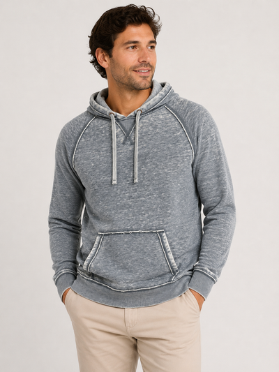 Wearing the Georg Roth Malibu Hoodie PHW by Georg Roth—a blue-gray cotton hoodie with white drawstrings and a front pocket—a man stands with his hands in the pockets of his beige pants, looking to the side, perfectly embodying casual comfort and dress-casual style.