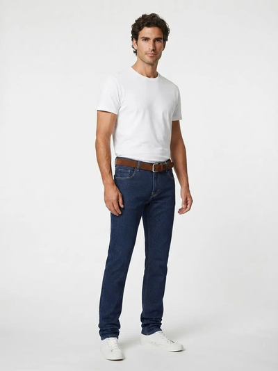 A man stands against a plain background, wearing a white t-shirt, Alberto Bill 1463 Raw Denim jeans by Alberto in a modern fit style, complemented by a brown belt and white sneakers.