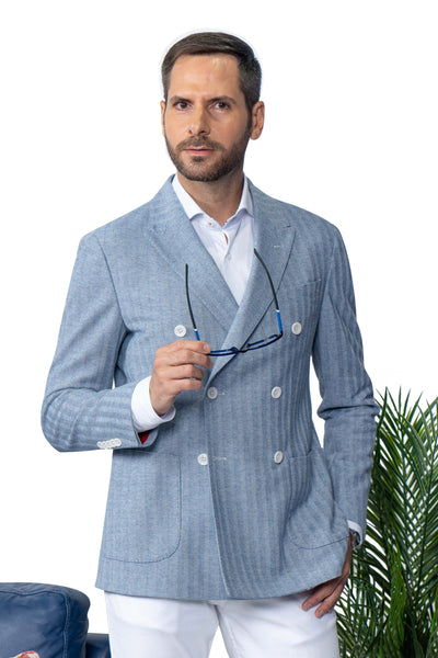 A man wears the 7 Downie St Dorval Sport Coat by 7 Downie St and white pants, holding glasses. He stands near a plant against a white background.
