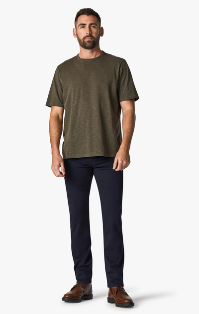 A man stands facing forward, wearing a short-sleeve olive green T-shirt, 34 Heritage Courage Twill Pant in a dark hue, and brown boots against a plain white background.