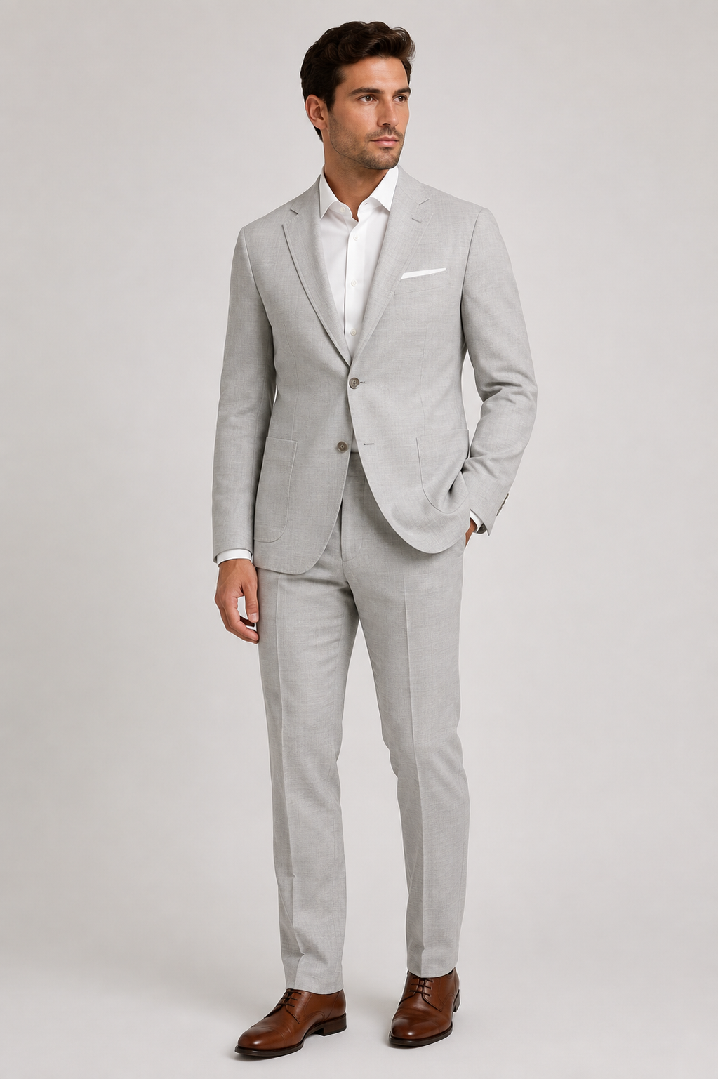 A man wearing the Paul Betenly OP Layton Slim Suit 1LH6120 in light gray, paired with a white dress shirt and brown dress shoes, stands confidently against a plain, light background.