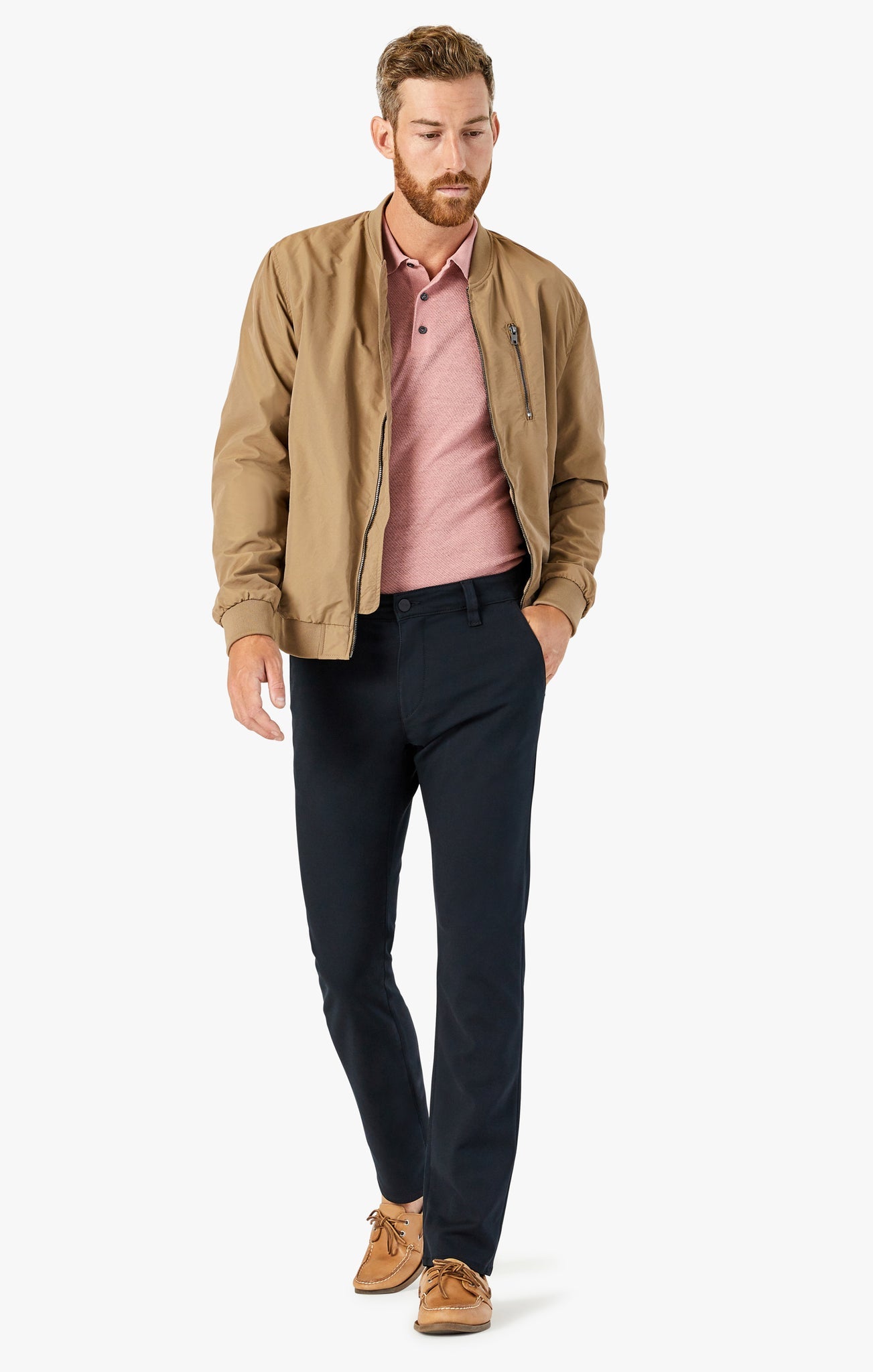 A man wearing a tan jacket, pink polo shirt, 34 Heritage Verona Pant High Flyer khaki trousers, and tan boat shoes walks forward with his left hand in his pocket against a plain white background.