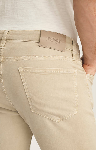 Close-up of the back pocket and waistband of 34 Heritage Courage Jeans Urban in beige, featuring a tan leather 34 Heritage patch, worn by a person in a white shirt.