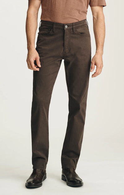 A man wearing a brown short-sleeve shirt, 34 Heritage Courage Twill Pant by 34 Heritage in dark brown, and dark brown boots stands against a plain light background.
