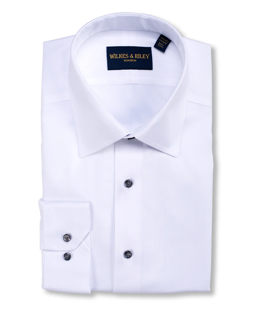 The Wilkes & Riley Herringbone B1515 is a neatly folded white spread collar dress shirt with black buttons, featuring premium Supima cotton and the Wilkes & Riley label inside the collar.