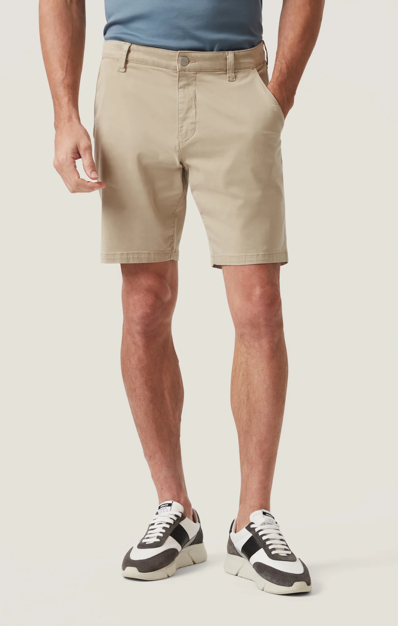 A man stands with his hands in his pockets against a plain background, wearing a blue t-shirt, 34 Heritage Arizona Twill Short by 34 Heritage, and black-and-white sneakers.