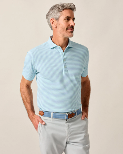 A man wears the Johnnie-O Performance Polo - Stetsons by Johnnie-O, styled with light gray pants and a blue belt with brown accents, posing hands-in-pockets against a neutral background.
