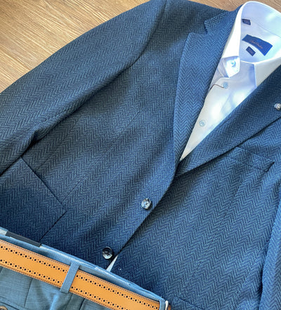 A Jack Victor Sport Coat C01252 in navy herringbone, paired with a white dress shirt, gray trousers, and a tan leather belt from the Comfortwear Collection, arranged on a wooden surface.