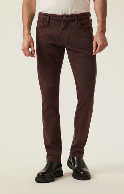 A man stands against a plain light background wearing 34 Heritage Courage Diagonal Pants in coffee, paired with a white shirt and black leather boots.