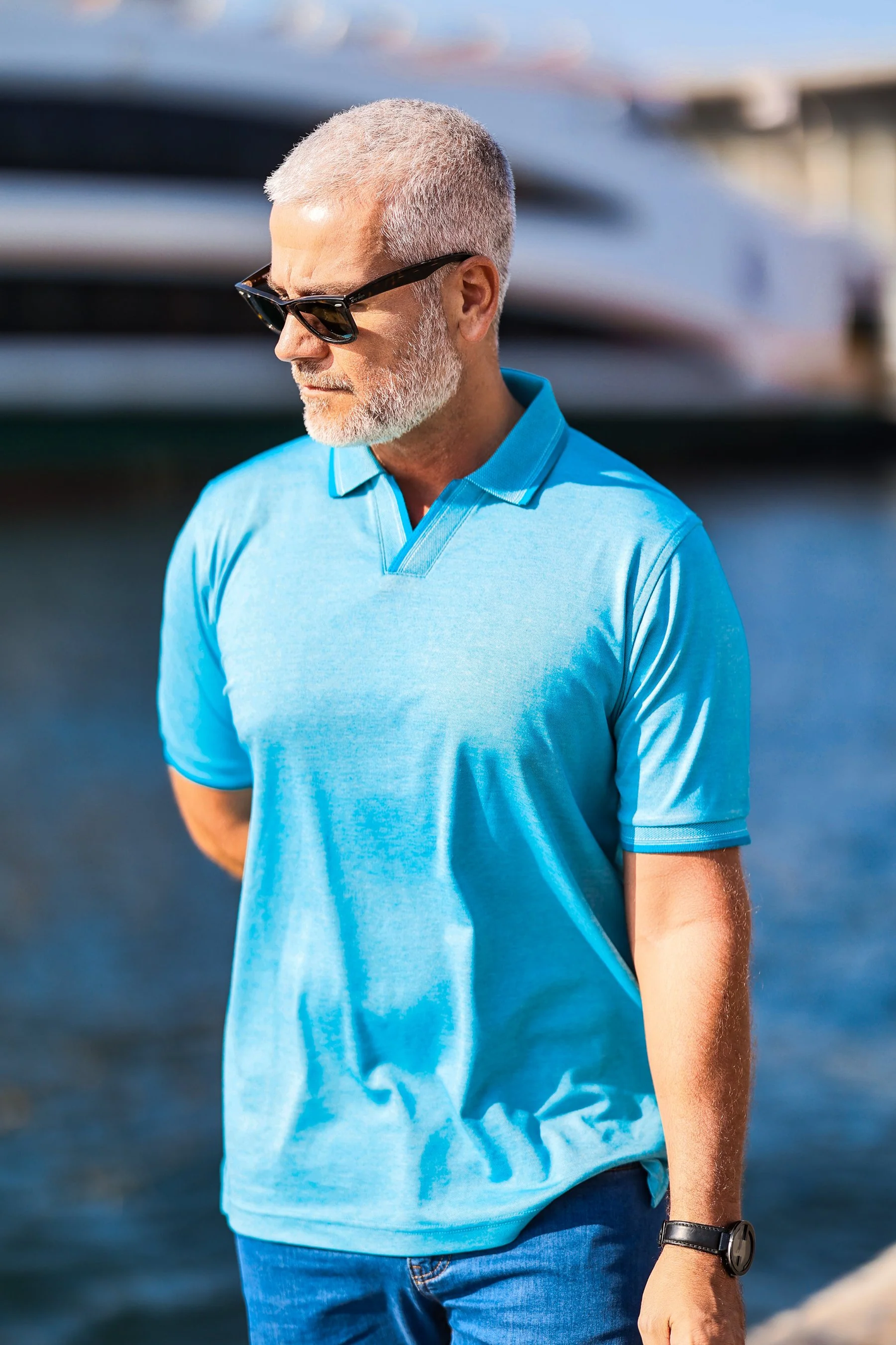 A man with gray hair and beard, wearing sunglasses and a Marcello Polo P-229 by Marcello in light blue royal oxford, stands near the water with a yacht in the background.