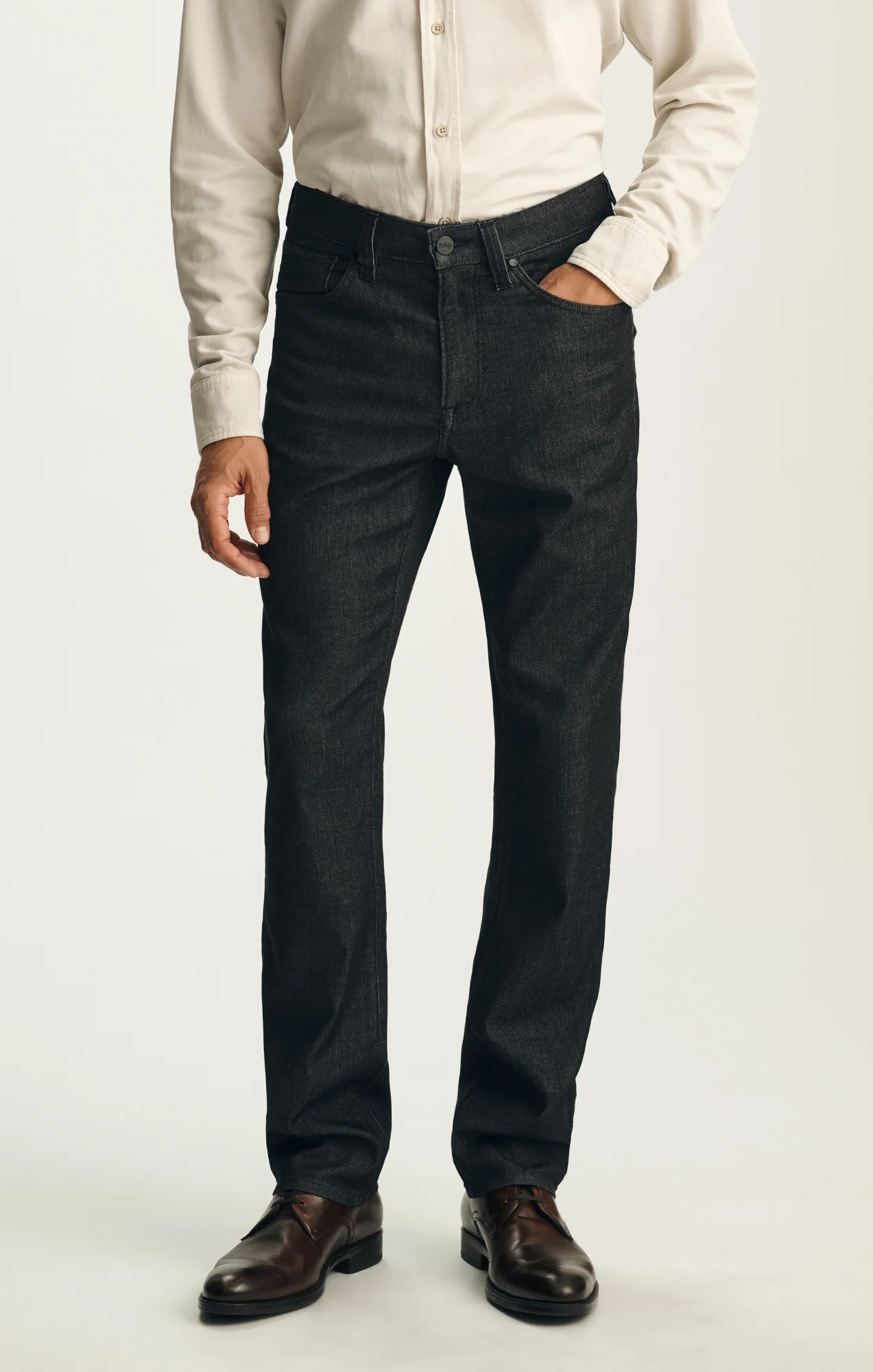 A person stands against a plain background with one hand in their pocket, dressed in a beige button-up shirt, brown dress shoes, and 34 Heritage Charisma Sporty Big&Tall relaxed straight leg trousers by 34 Heritage.