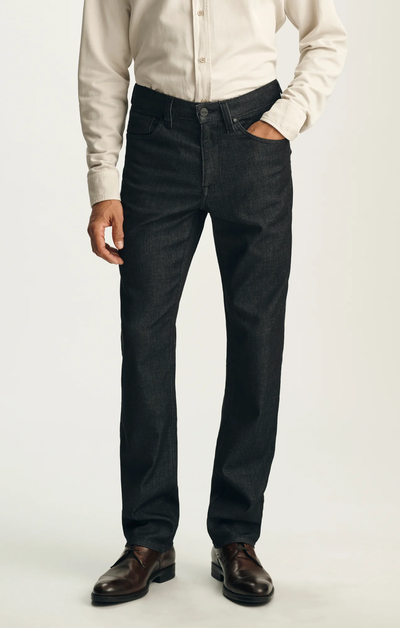 A person stands against a plain background with one hand in their pocket, dressed in a beige button-up shirt, brown dress shoes, and 34 Heritage Charisma Sporty Big&Tall relaxed straight leg trousers by 34 Heritage.