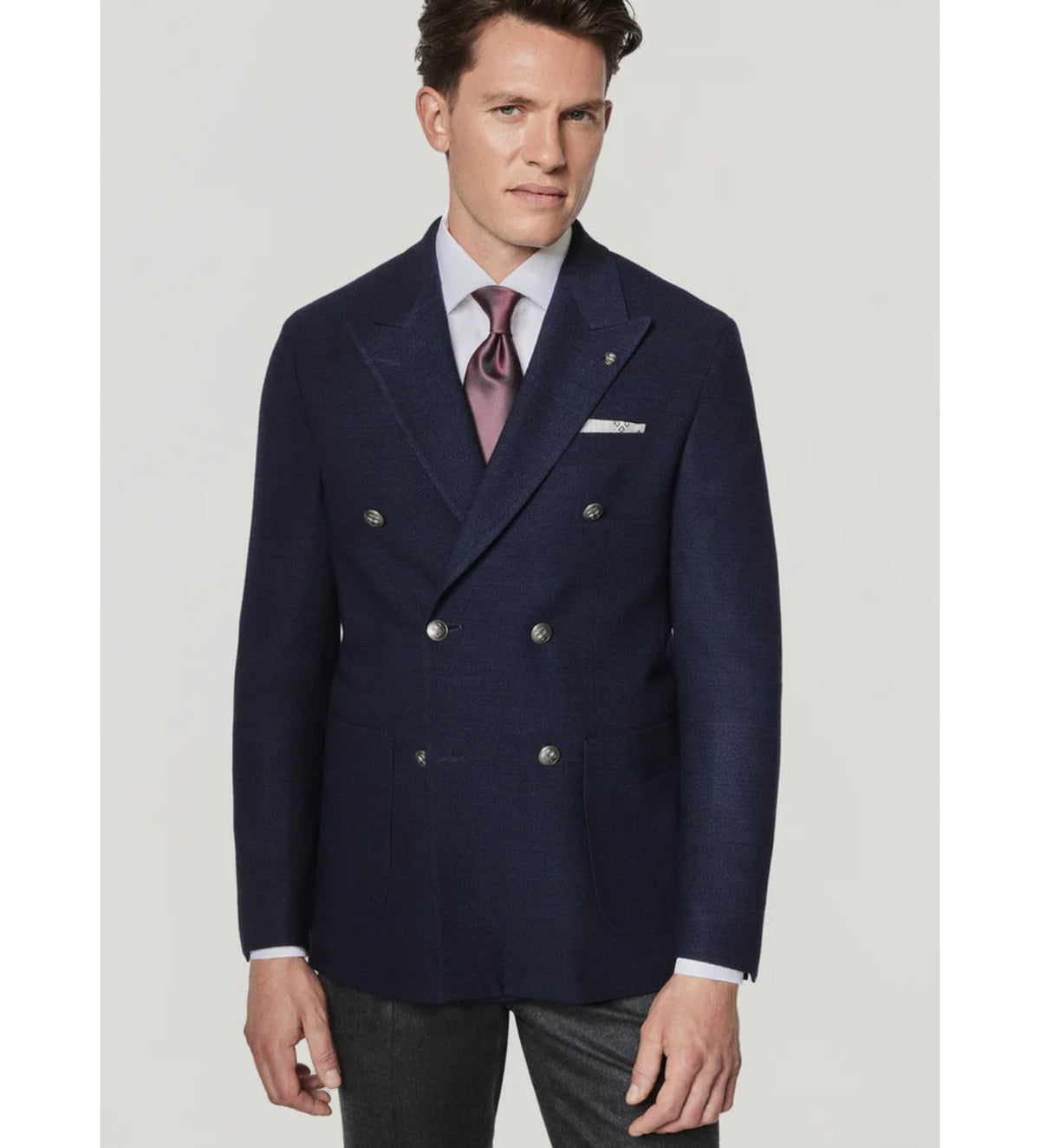 A man wearing the Jack Victor Hampton SC C1273 tailored-fit navy double-breasted sport coat, crafted from 100% wool by Jack Victor, paired with a lavender dress shirt, pink tie, and gray trousers, stands against a plain light background.