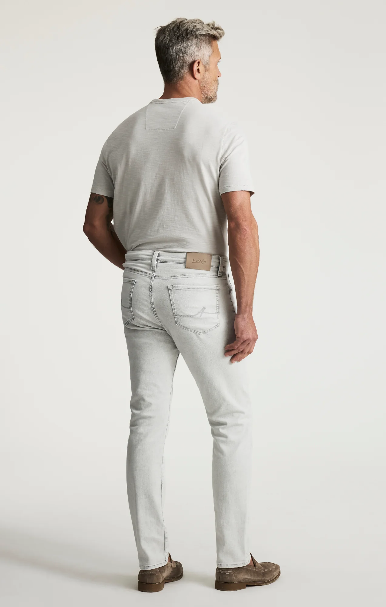 A man stands facing away, wearing a light gray t-shirt, 34 Heritage Courage Jeans Organic Ultra by 34 Heritage, and brown shoes against a plain white background.