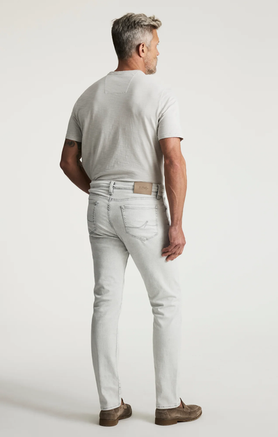 A man stands facing away, wearing a light gray t-shirt, 34 Heritage Courage Jeans Organic Ultra by 34 Heritage, and brown shoes against a plain white background.
