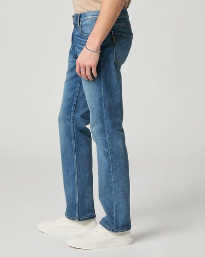 A person wears Paige Federal Straight Slim Jeans by Paige in a medium wash, styled with a beige shirt and white sneakers, standing against a plain light background.