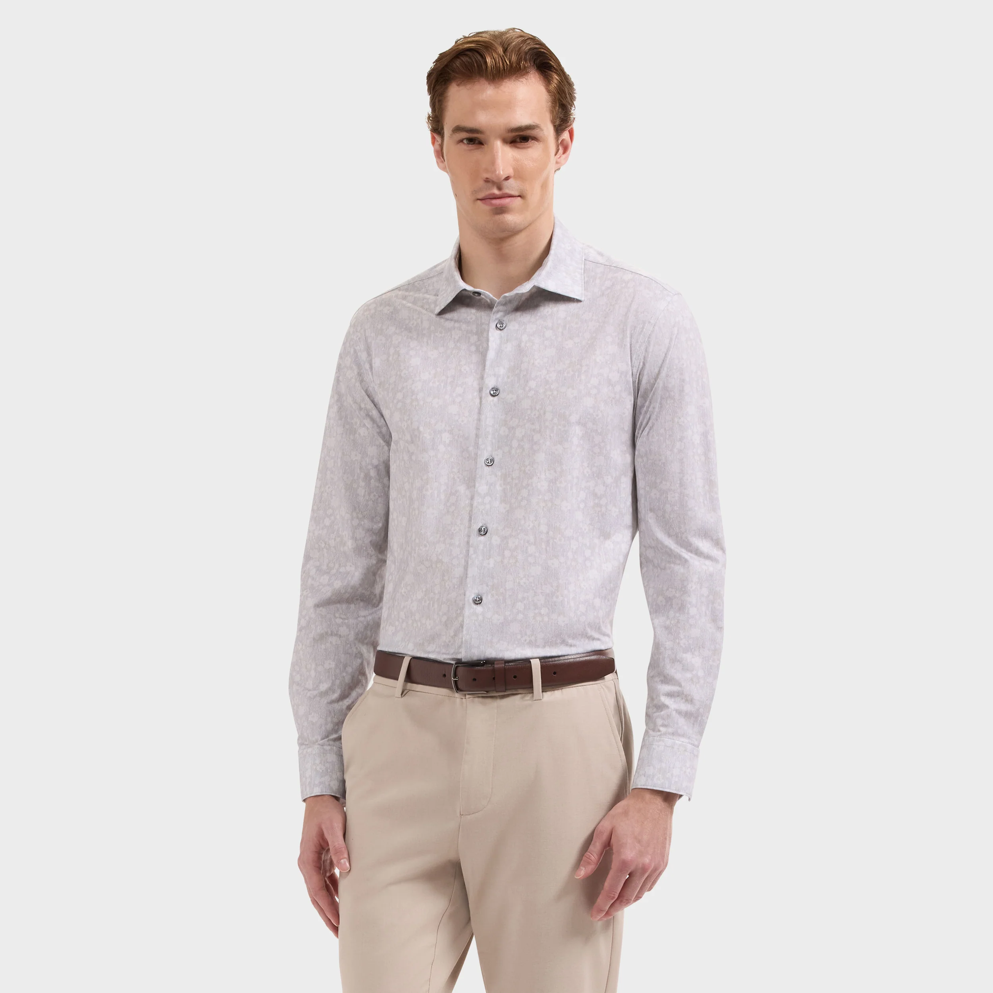 A man is wearing the Bugatchi James Floral Sport Shirt (KF9142K60) by Bugatchi, crafted from wrinkle-resistant OoohCotton fabric with 8-way stretch in a light grey patterned design. He pairs it with light beige pants and a brown belt, standing against a plain white background.