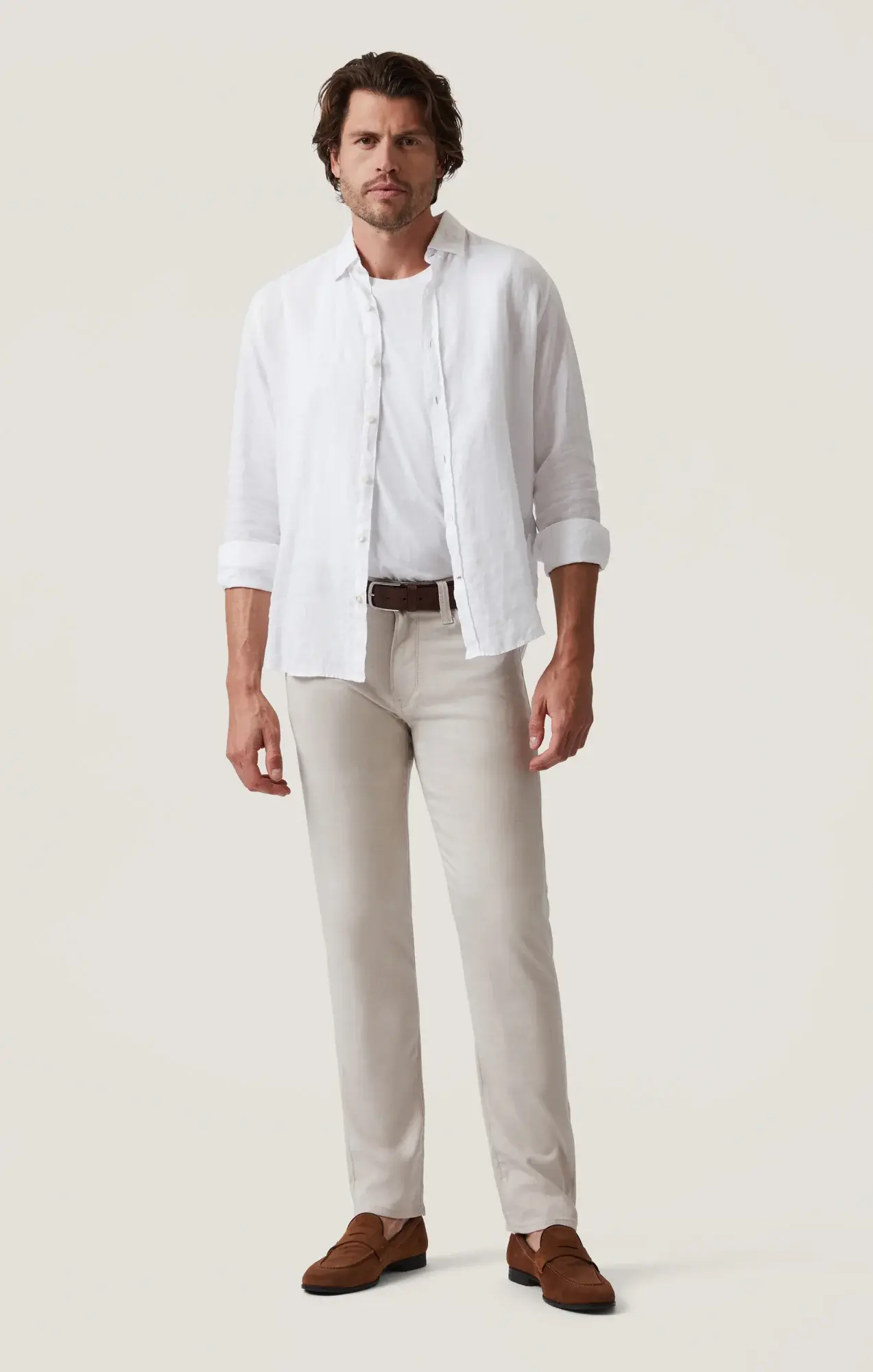 A man stands against a plain background, dressed in a white shirt over a white t-shirt, and beige 34 Heritage Courage Cross Twill straight-leg pants by 34 Heritage. He completes the look with a brown belt and brown loafers.