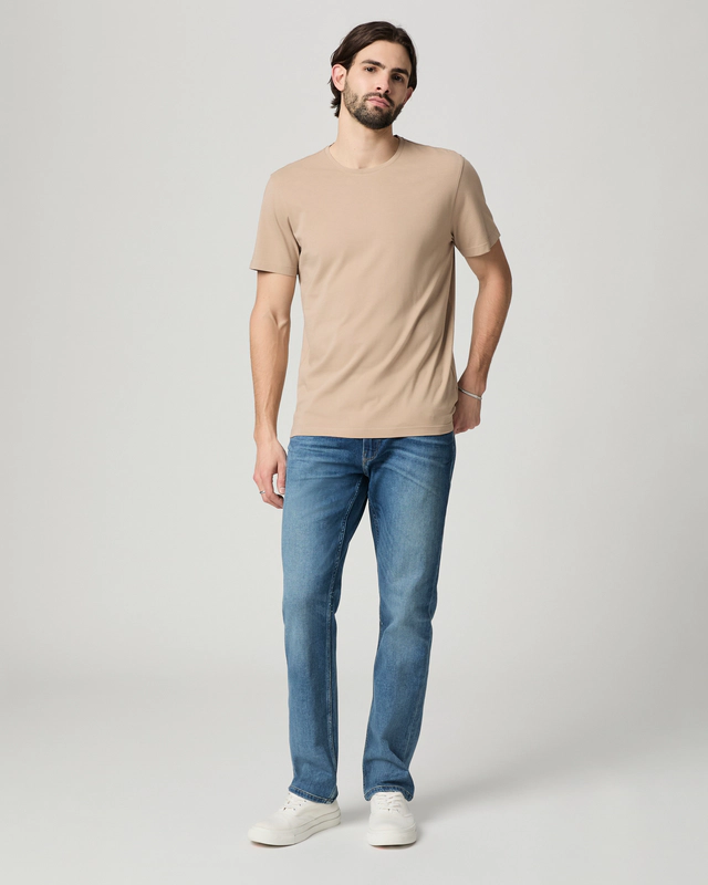 A man stands against a plain background wearing a beige t-shirt, white sneakers, and Paige Federal Straight Slim Jeans in a medium wash.