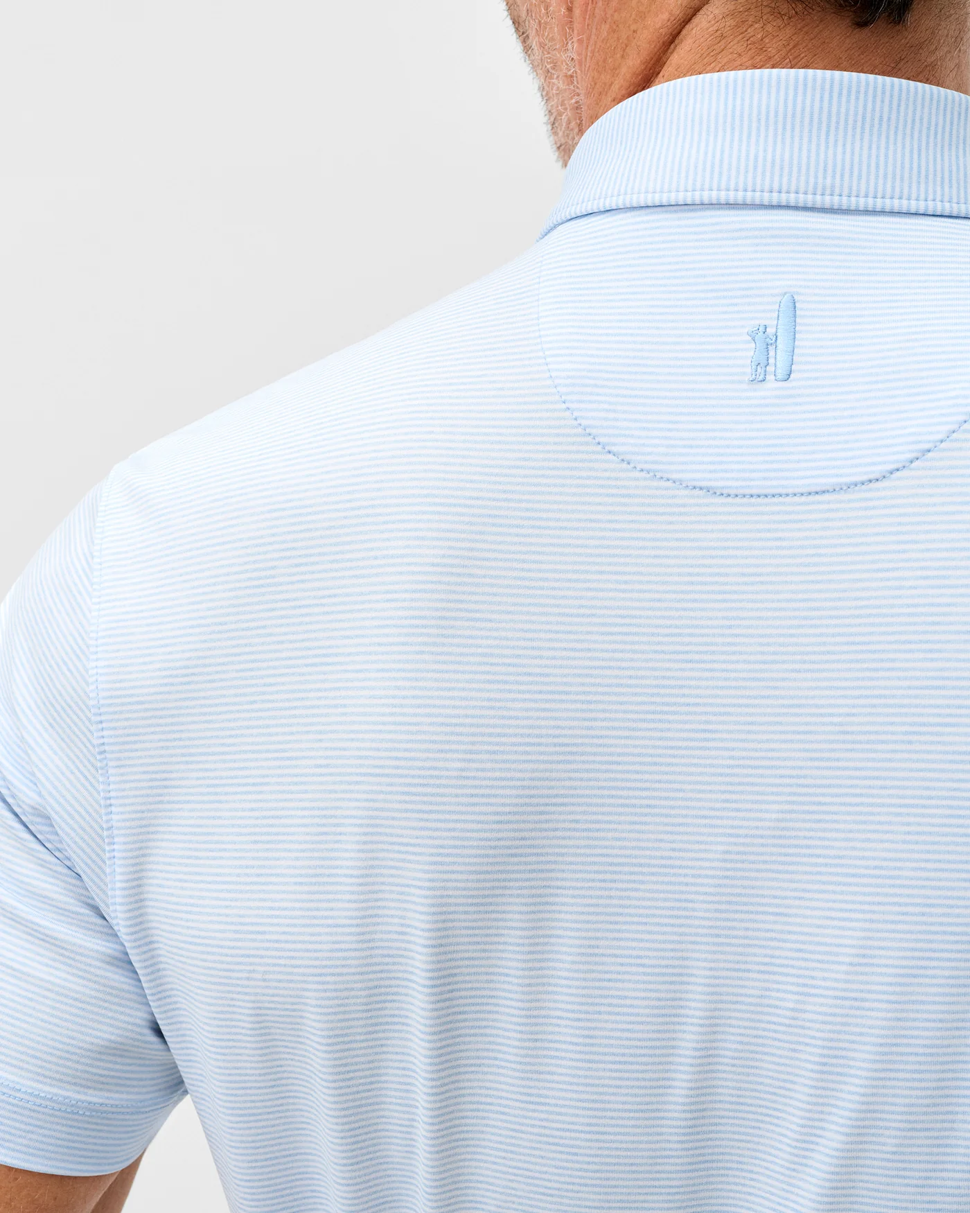 A person wearing the Johnnie-O Lyndonn Stripe Performance Knit Polo, a light blue, casual short-sleeve striped shirt by Johnnie-O with a small embroidered logo near the collar on the upper back.