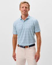 A man stands facing forward against a white background, wearing the Johnnie-O Charmer Jersey Polo in light blue with a pattern, paired with beige pants and a dark belt.