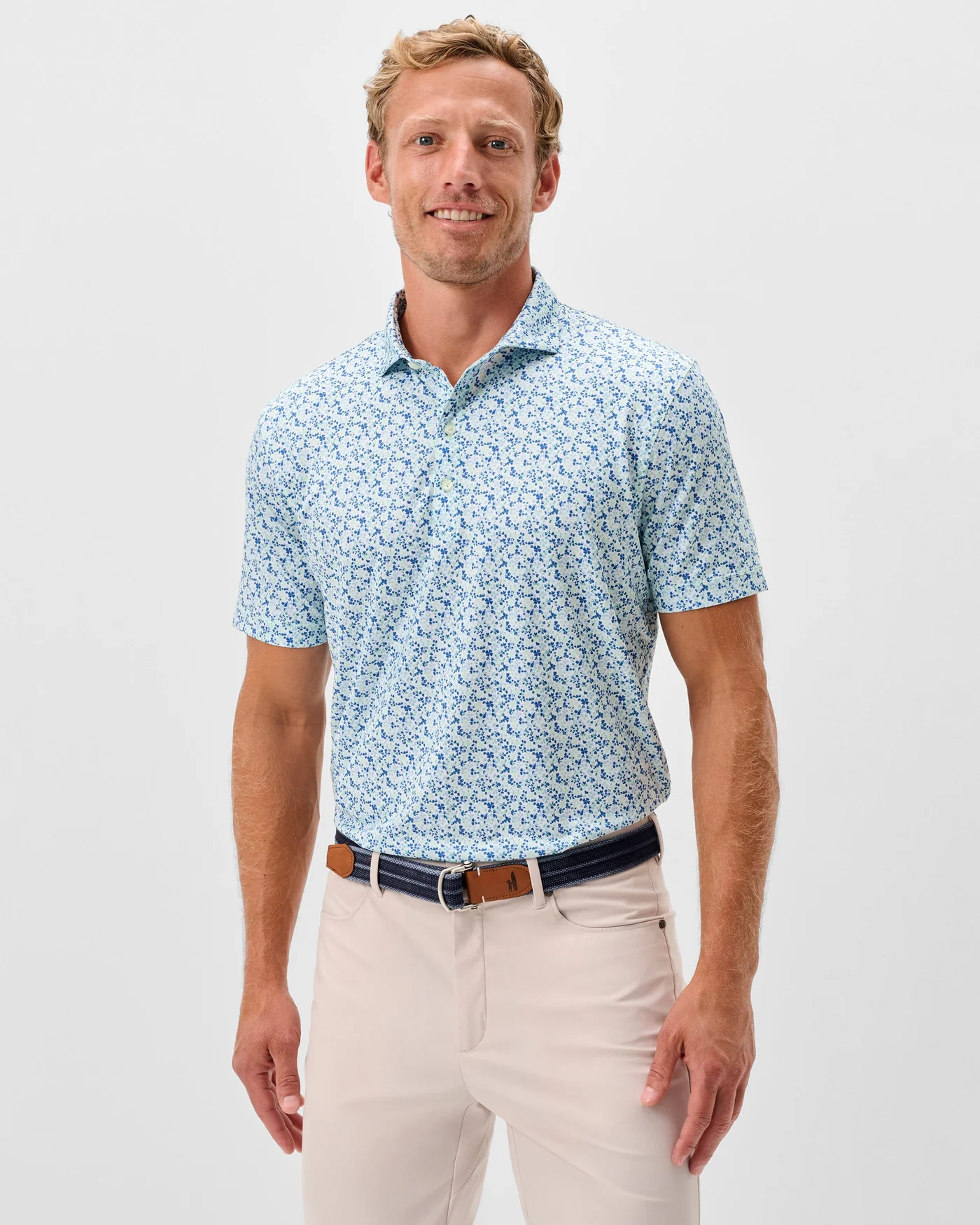 A man stands facing forward against a white background, wearing the Johnnie-O Charmer Jersey Polo in light blue with a pattern, paired with beige pants and a dark belt.