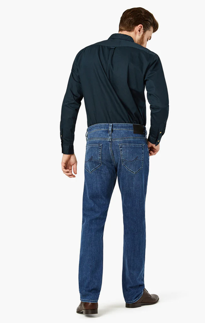 A man stands facing away against a plain white background, wearing a dark long-sleeve shirt, brown shoes, and 34 Heritage Charisma Mid Urban premium stretch denim straight leg jeans by 34 Heritage.