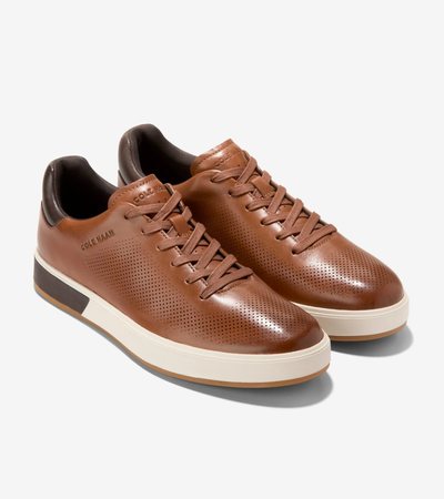 The Cole Haan GrandPro Angleace Sneakers by Cole Haan are brown leather low-top sneakers with a minimalist design, perforated sides, leather upper, white rubber soles, and brown laces, shown on a white background.