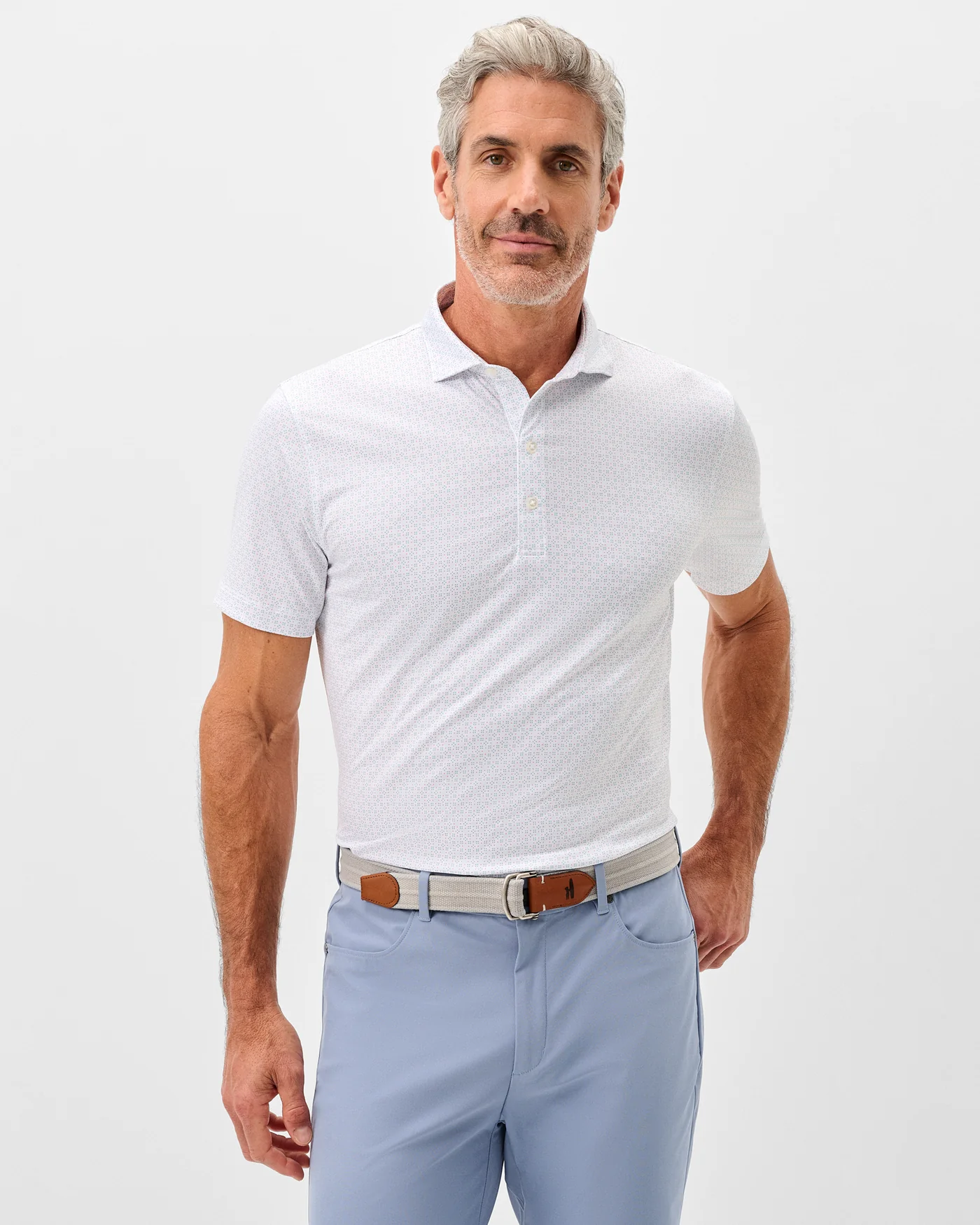 A man with gray hair and a beard wears the Johnnie-O Torres Performance Big & Tall Polo with light blue pants and a belt, standing with one hand in his pocket against a plain light background.