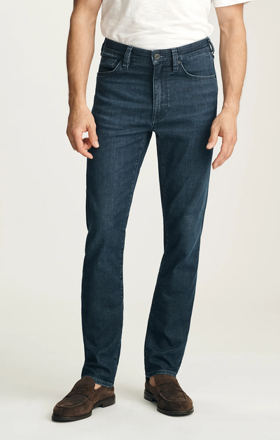 A person wearing a white shirt, 34 Heritage Courage Sporty Jeans in dark blue denim enhanced with Thermocool fiber technology, and brown loafers stands against a plain background.