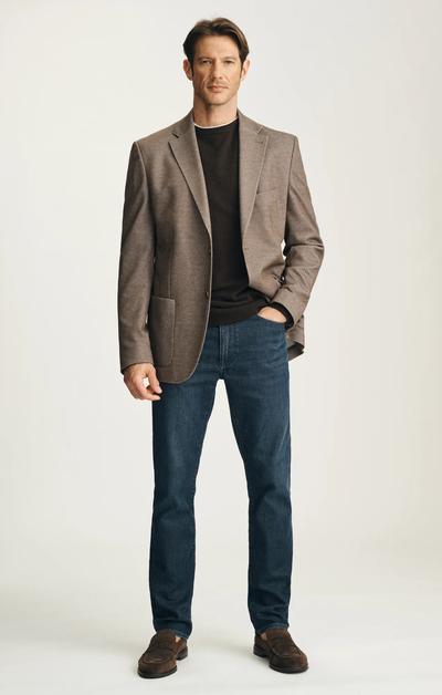 A man stands against a plain background, wearing a brown blazer, dark sweater, 34 Heritage Courage Sporty Jeans in dark blue by 34 Heritage, and brown shoes, with one hand in his pocket.
