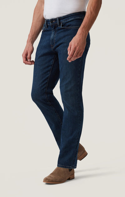 Wearing 34 Heritage Mid Comfort higher-rise, straight leg jeans by 34 Heritage with a classic five-pocket style, a white top, and brown suede boots, the person stands against a plain light background.